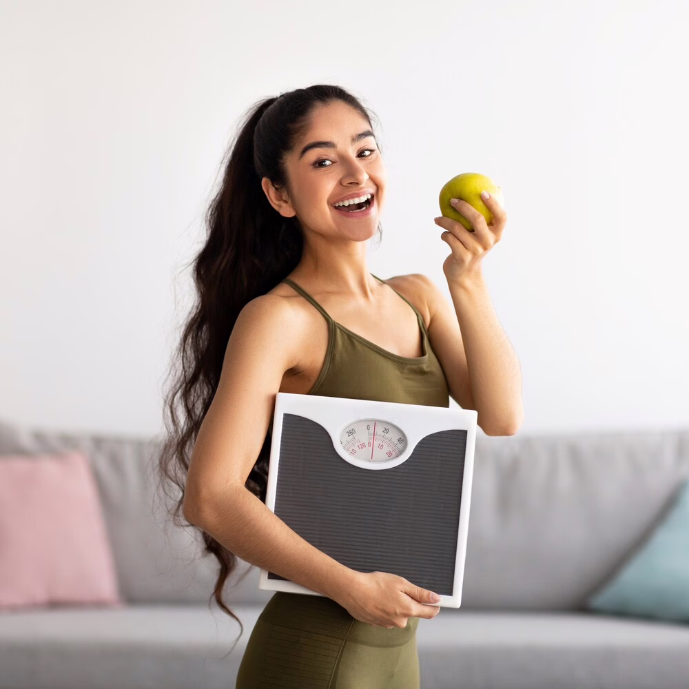 Portrait of millennial Indian lady holding scales and eating apple, choosing healthy diet, panorama. Young Asian woman preferring wholesome nutrition. Wellbeing and weight loss concept. How to weight loss fast?